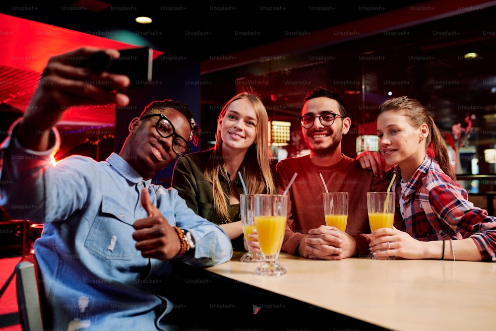 Joven africano con un teléfono inteligente que muestra el pulgar hacia arriba mientras se hace selfie con sus amigos en la mesa en un café en un centro de ocio
