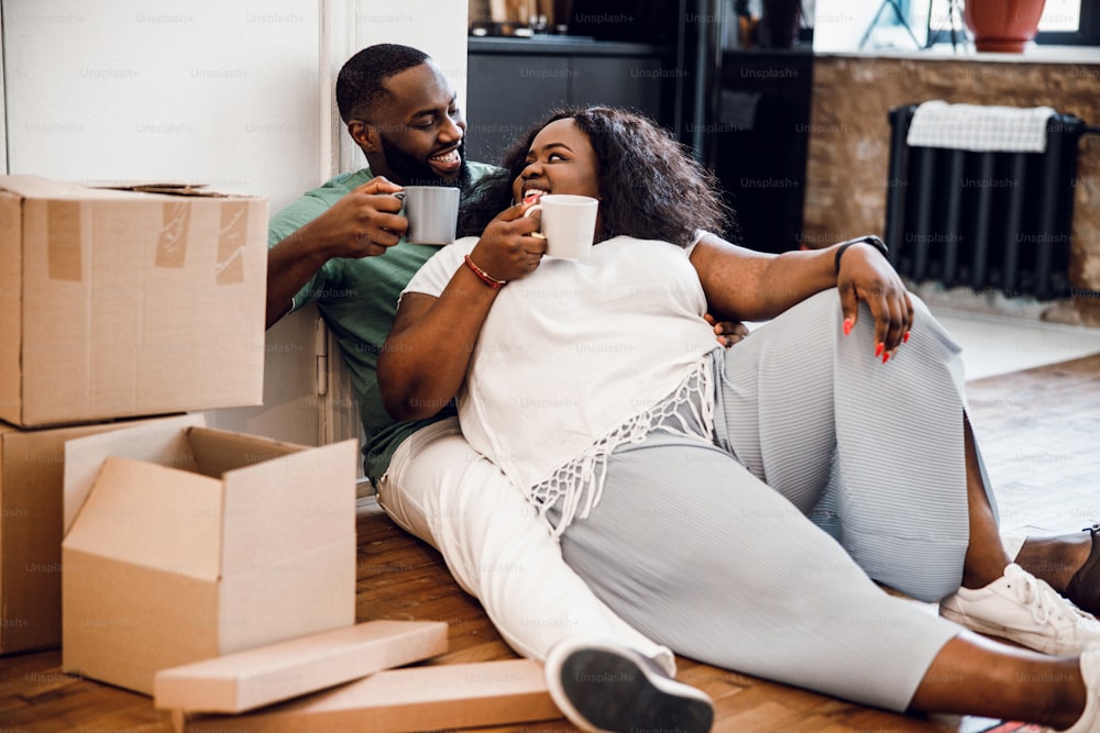 Mirthful man and woman sitting on the floor near boxes and drinking coffee on the moving day