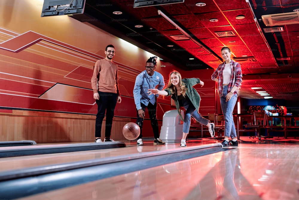 Pretty young blond female throwing bowling ball on track while standing among her intercultural friends during play