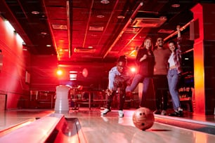 African guy bending by bowling alley while throwing ball on background of his friends with raised arms expressing gladness