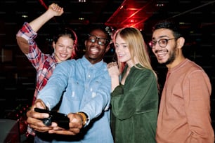 Four joyful young men and women in casualwear looking at smartphone camera and making selfie while enjoying party in club