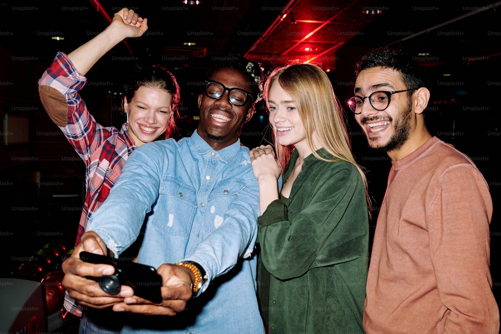Cuatro hombres y mujeres jóvenes alegres con ropa casual mirando la cámara del teléfono inteligente y haciendo selfies mientras disfrutan de la fiesta en el club
