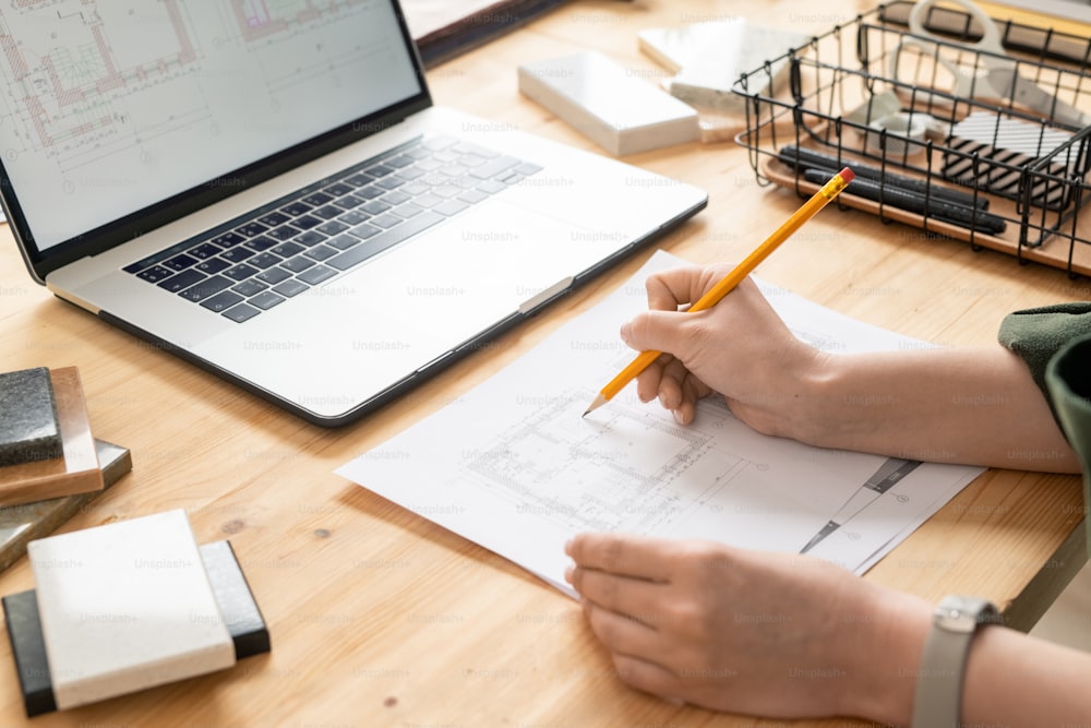 Hand of young female architect with pencil over peper with sketch of house during work over new project in front of laptop