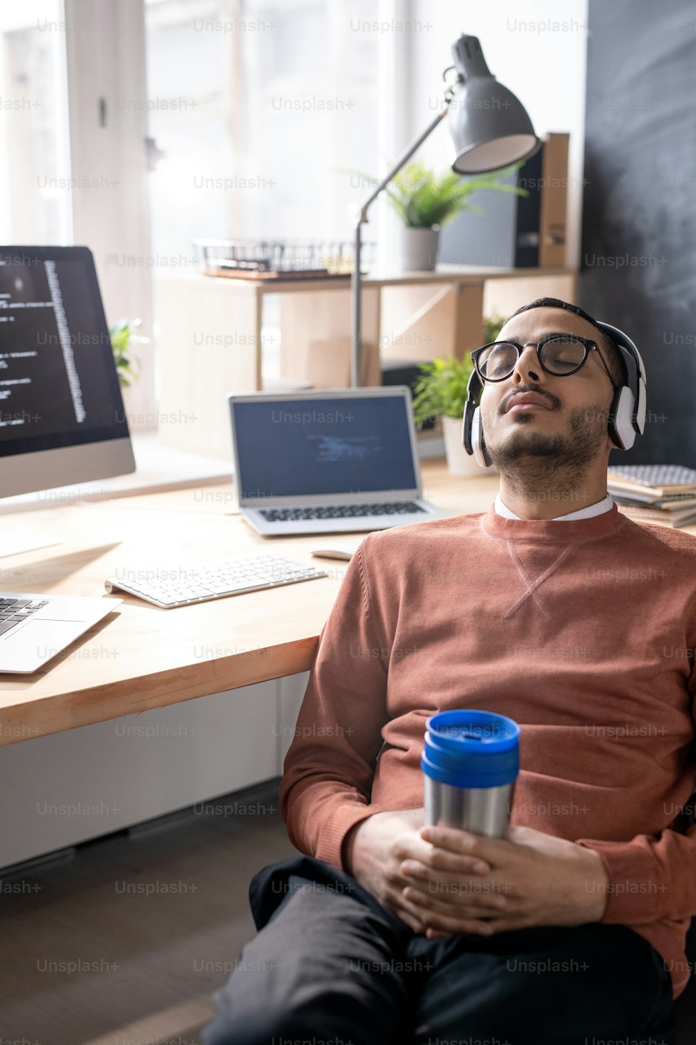 Exhausted young Arabian coder in headphones and glasses sleeping with thermos cup in hands at workplace