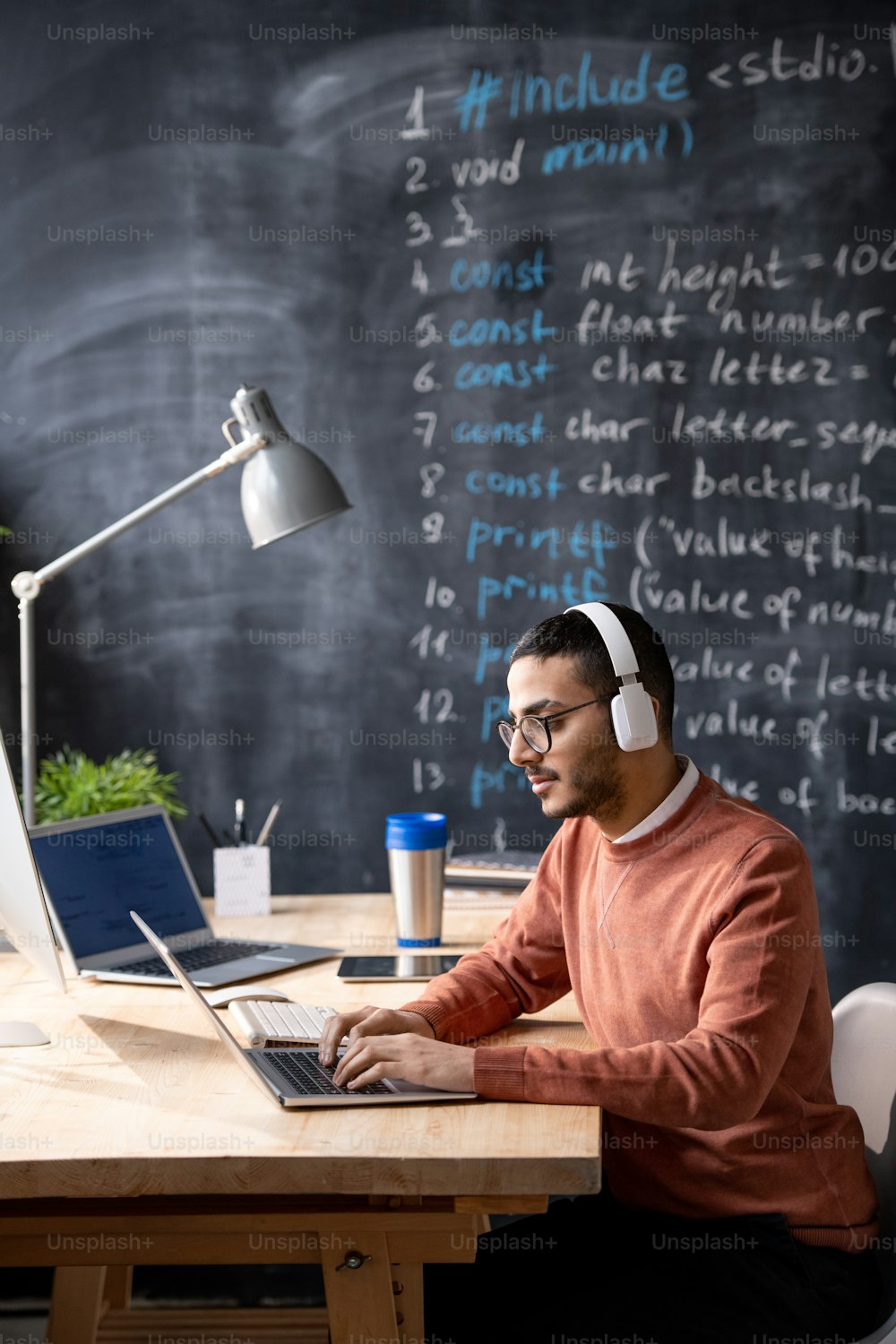 Joven programador árabe centrado en el código web sentado en una mesa de madera y escribiendo en la computadora portátil mientras escucha audio en auriculares