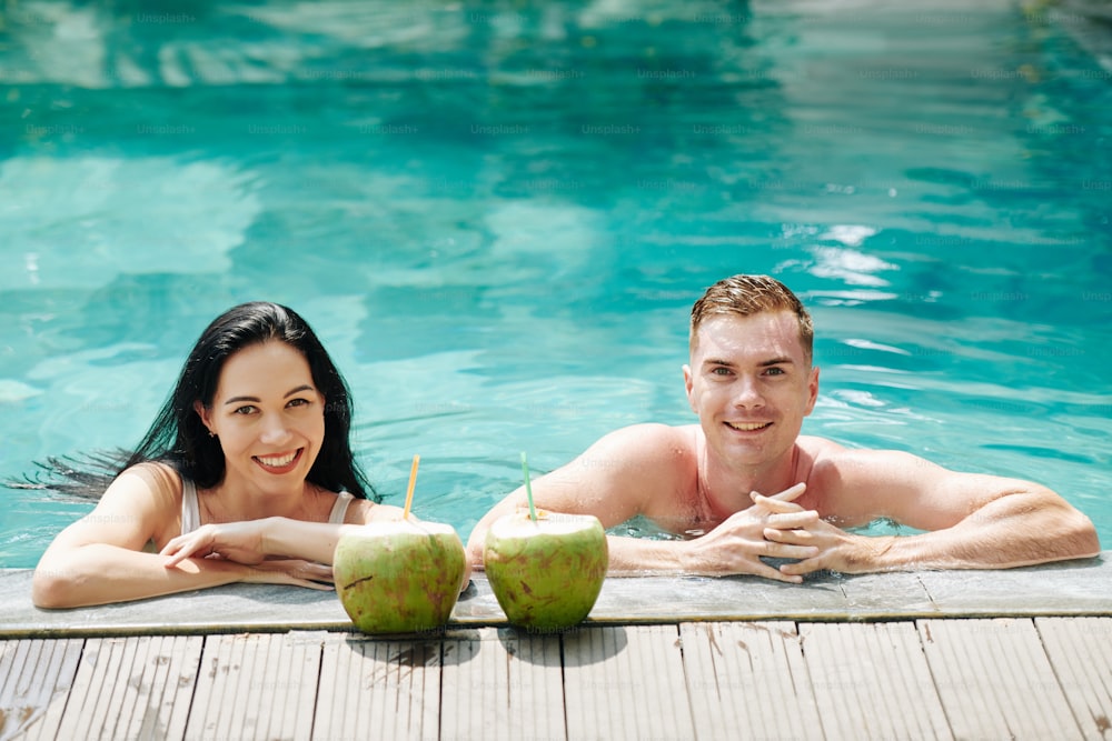 Cheerful young couple with beautiful smiles leaning on edge of swimming pool and looking at camera