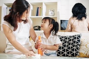 Mãe ensinando a filha pequena a limpar a superfície da mesa com spray desinfetante