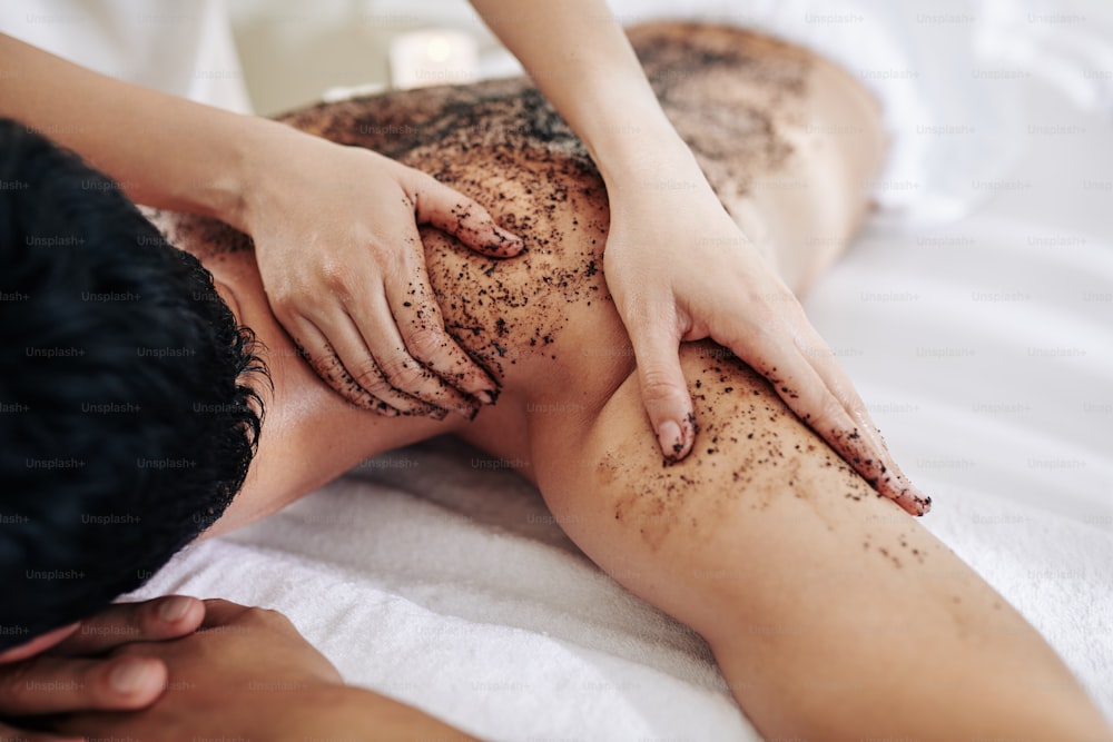 Cosmetologist applying natural apricot scrub on back and shoulders of man relaxing in spa salon
