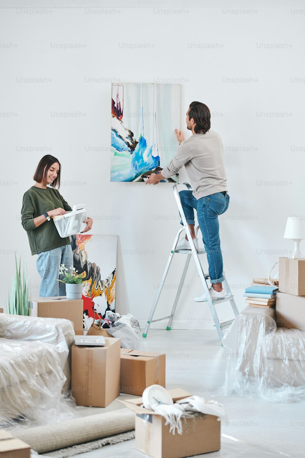 Joven con ropa casual de pie en una escalera junto a la pared y colgando pintura abstracta mientras su esposa mira la imagen en el marco
