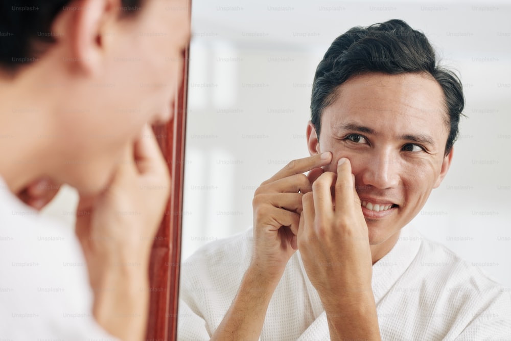 Smiling mixed-race man looking at mirror and squeezing pimples