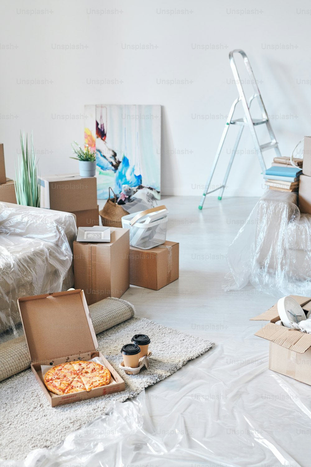 Open box with delivered appetizing pizza and two glasses of coffee on the floor by couch surrounded by packed boxes and other stuff
