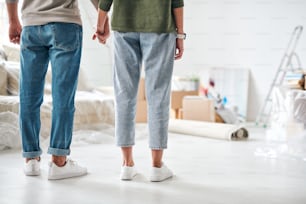 Back view of affectionate young couple in jeans and pullovers holding by hands while standing in living-room of new house or flat