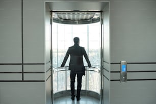 Rückansicht eines jungen, eleganten Geschäftsmannes in formeller Kleidung, der im Aufzug mit offener Tür steht, während er die Pause im Bürozentrum genießt