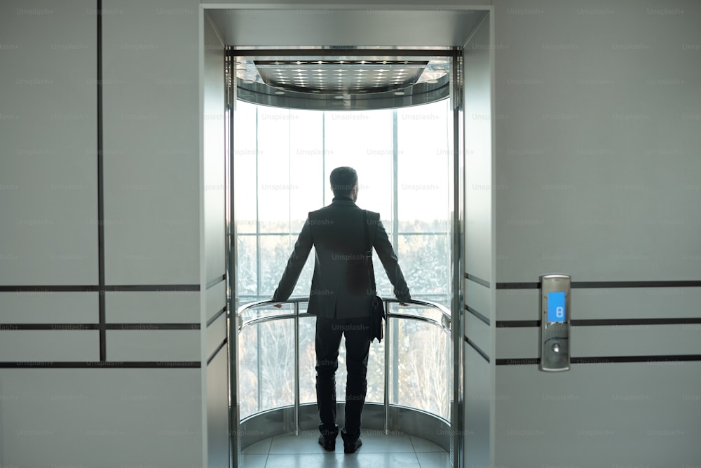 オフィスセンターで休憩を楽しみながら、ドアを開けてエレベーターに立つフォーマルウェアを着た若い優雅なビジネスマンの背面図