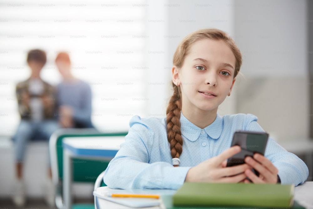 Porträt eines süßen blonden Mädchens, das ein Smartphone hält, während es am Schreibtisch im Klassenzimmer der Schule sitzt und in die Kamera schaut, Kopierraum
