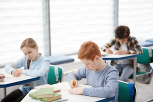 Vista ad alto angolo su un gruppo multietnico di bambini seduti ai banchi di scuola e scrivendo appunti di prova, spazio di copia