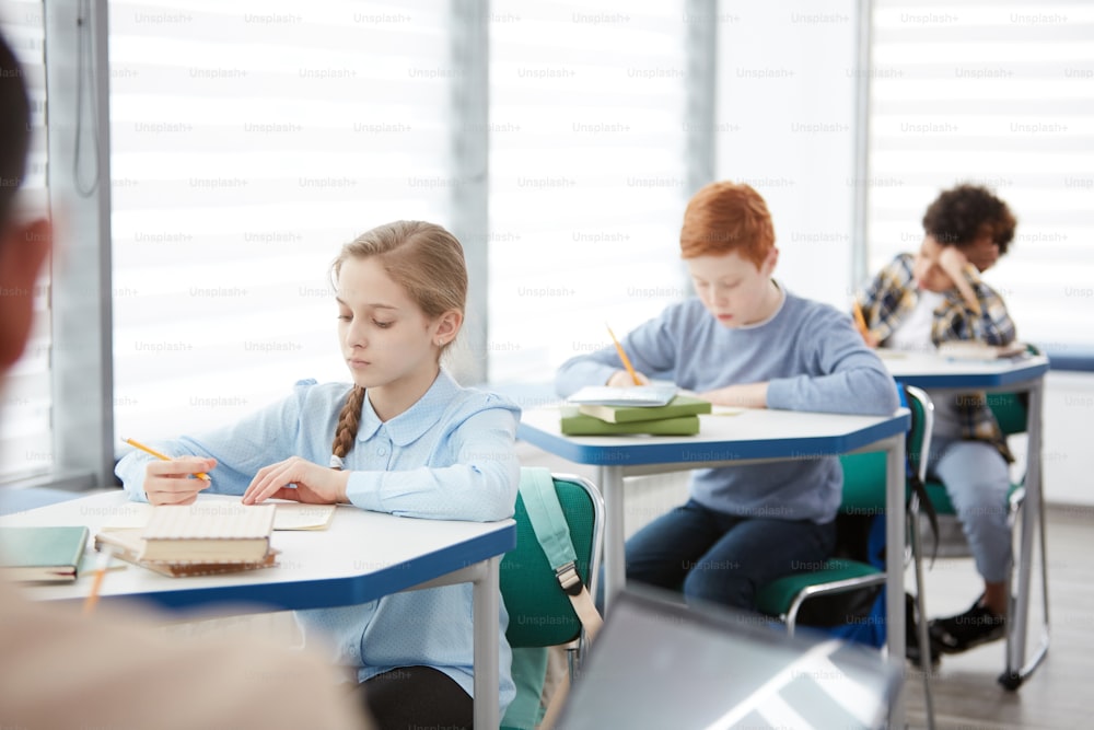 Vista laterale al gruppo multietnico di bambini seduti in fila alla scrivania nell'aula scolastica e scrivendo o facendo test, spazio di copia