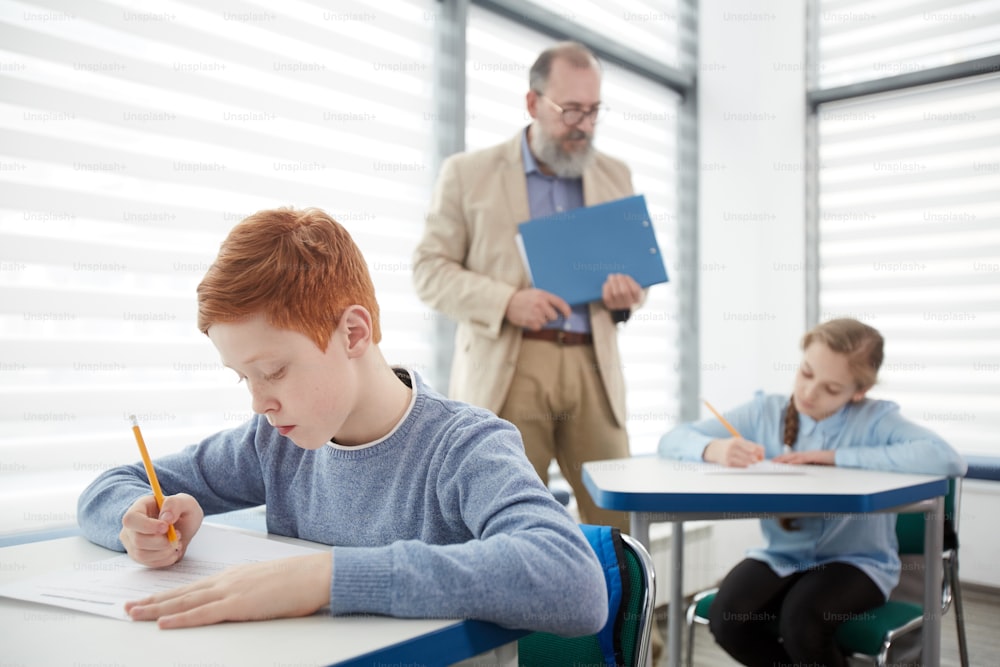 Group of children taking test with bearded mature teacher watching them, focus on red haired boy sitting at desk and writing in foreground, copy space