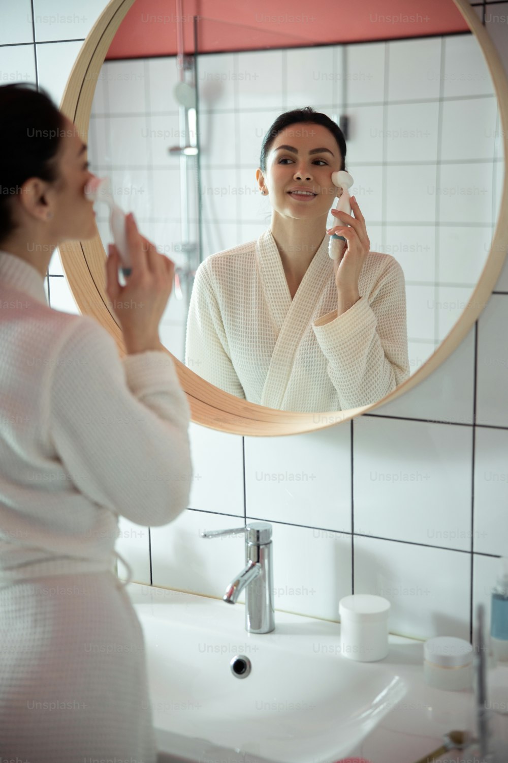 Relaxed woman is standing in front of mirror in bathroom and doing face massage with device