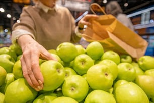 Mano della donna anziana con il sacchetto di carta che prende la mela fresca della nonna smith mentre si trova vicino all'esposizione della frutta nel supermercato contemporaneo