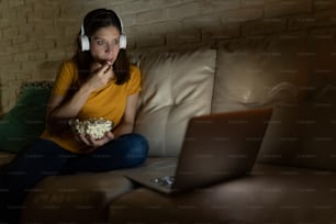 Femme choquée regardant un film à suspense sur un ordinateur portable la nuit alors qu’elle est assise dans un canapé à la maison
