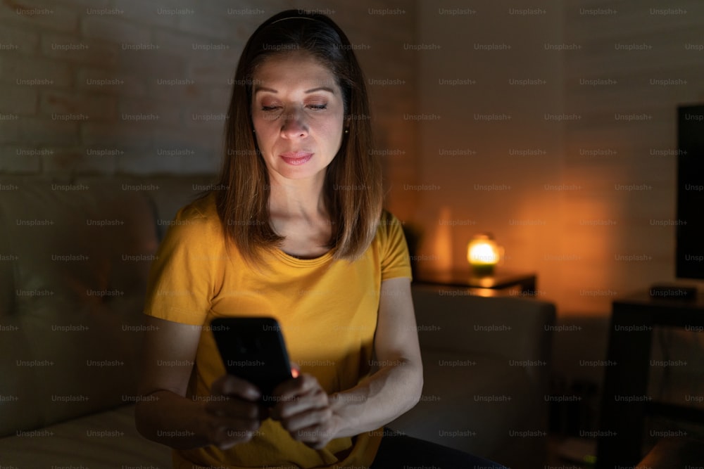 Mulher de boa aparência na casa dos 40 anos usando um smartphone para enviar mensagens de texto e verificar as notícias enquanto está sentada na sala de estar à noite
