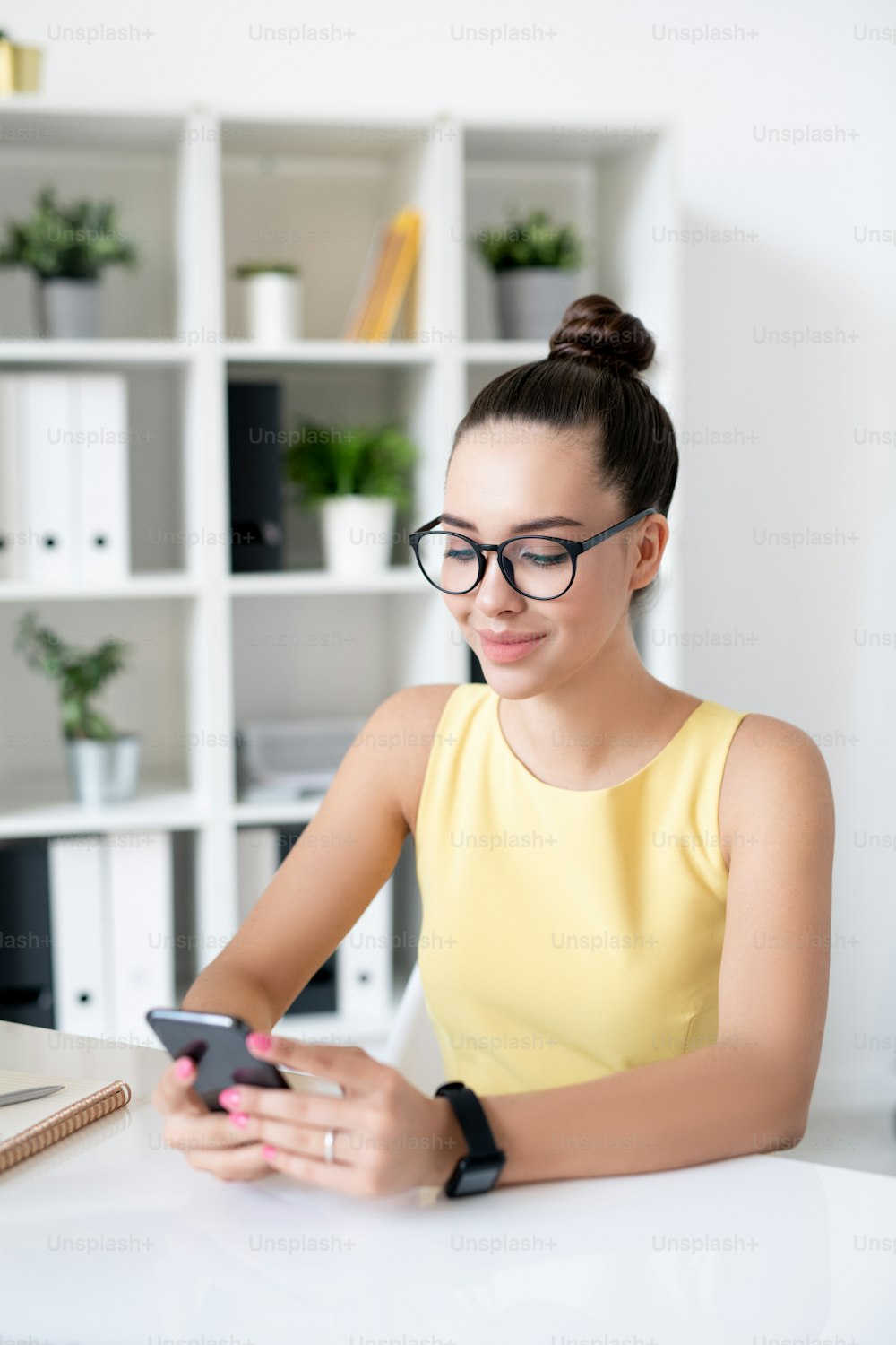 Lächelnde attraktive junge Geschäftsfrau mit Haarknoten, die am Tisch sitzt und das Smartphone benutzt, während sie in der Pause in den sozialen Medien surft