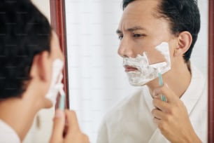 Homem sério franzindo a testa na cidade de waffle olhando para o espelho ao fazer a barba pela manhã