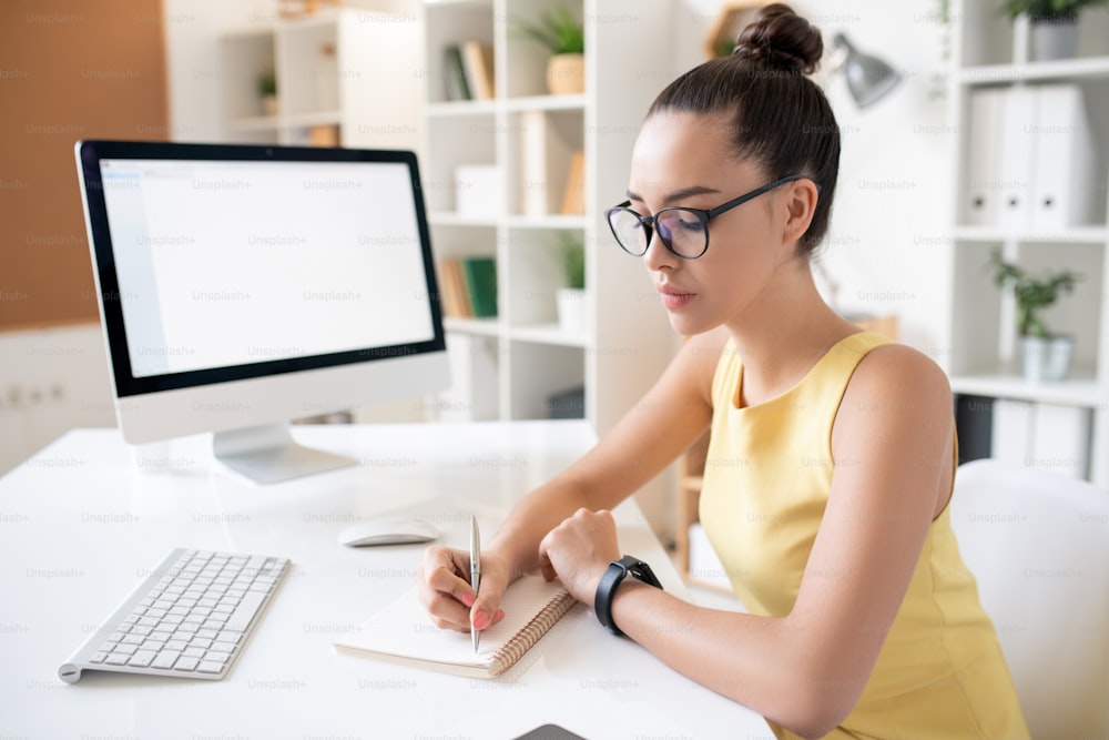 Mujer joven ocupada con moño de pelo sentada en el escritorio con la computadora y anotando en el diario mientras planifica las tareas en el trabajo
