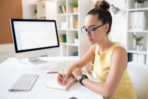 Giovane donna indaffarata con crocchia per capelli seduta alla scrivania con computer e annotando nel diario mentre pianifica le attività al lavoro