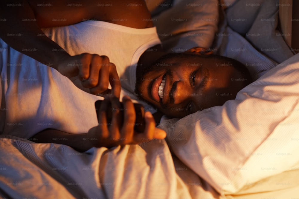 High angle portrait of smiling African-American man using smartphone in bed at night while texting girlfriend, copy space