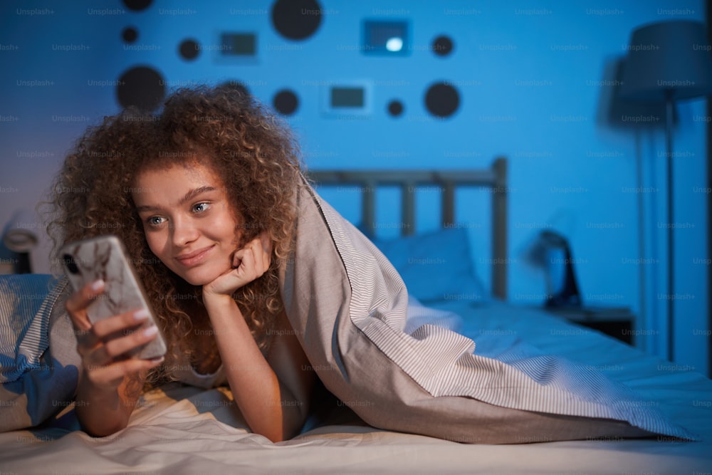 Retrato da jovem de cabelos cacheados olhando para a tela do smartphone e sorrindo enquanto deitada na cama à noite, espaço de cópia