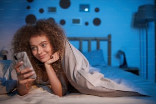 Retrato de una joven de cabello rizado mirando la pantalla del teléfono inteligente y sonriendo mientras está acostada en la cama por la noche, copie el espacio