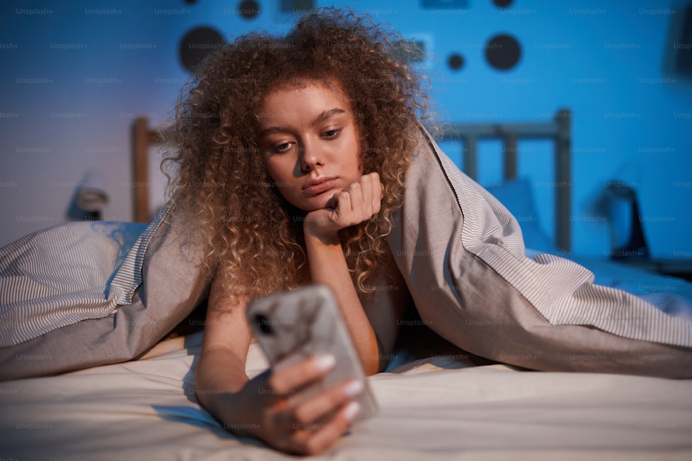 Retrato da vista frontal da jovem entediada usando o smartphone na cama à noite coberto por cobertor, espaço de cópia