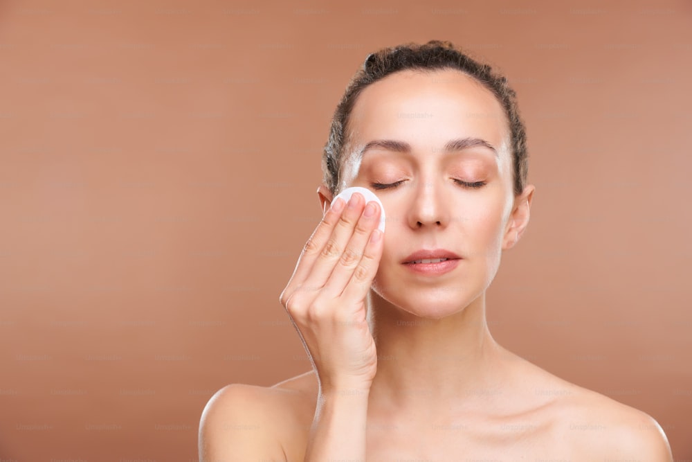Young beautiful lady cleaning her face with tonifying lotion in the morning or after removing makeup in the evening against brown background