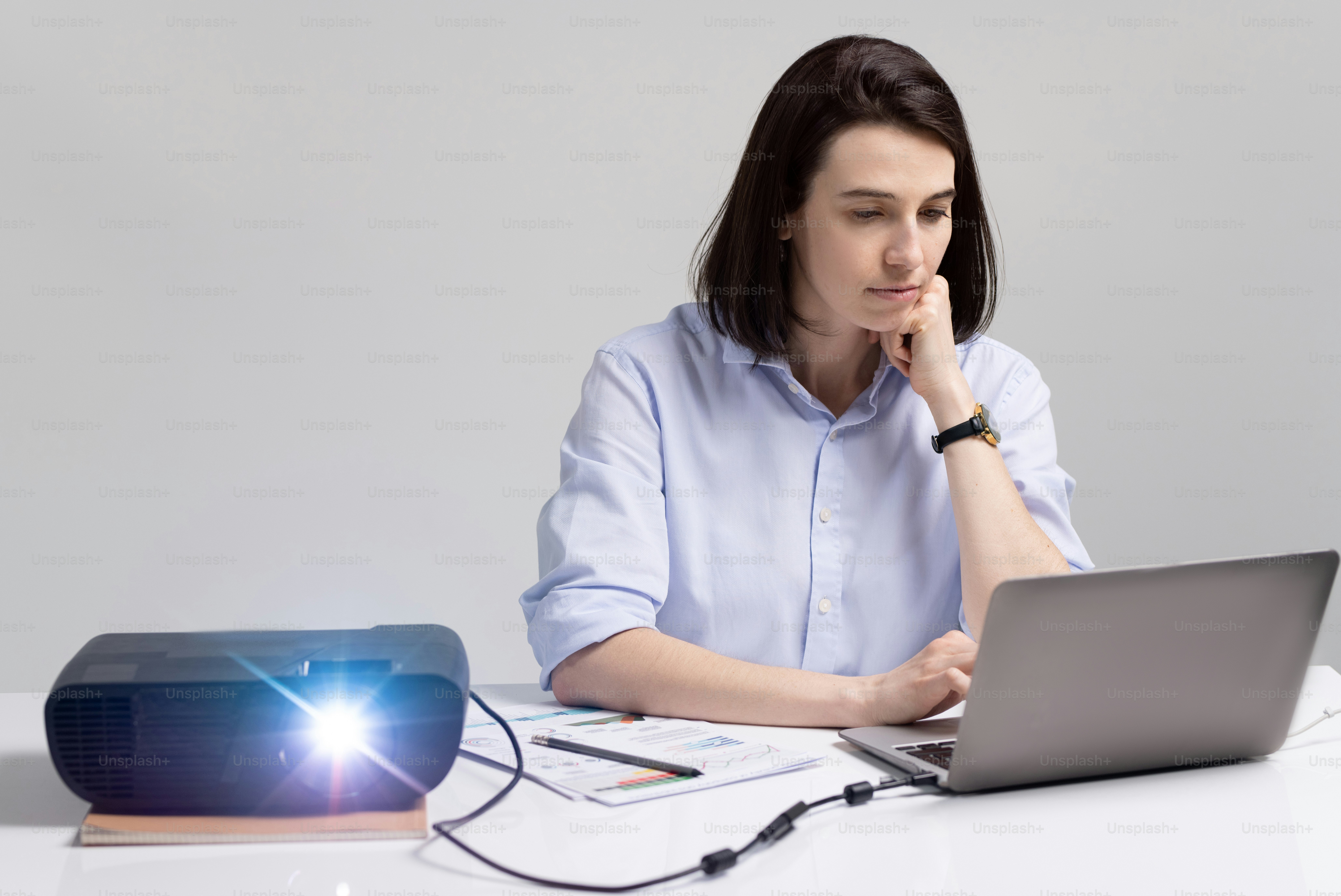 Pretty brunette student looking at laptop display while sitting by desk with switched on projector and preparing presentation before seminar