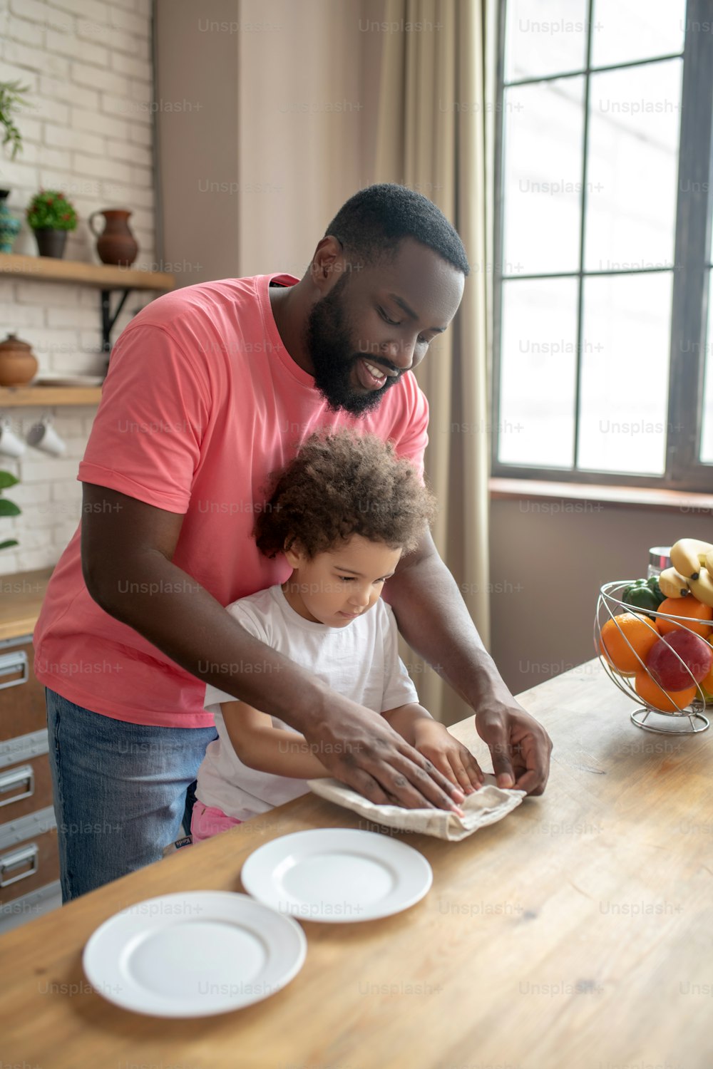Lets do it together. Tall dark-skinned man in a pink tshirt showing to his kid how to wipe the plate