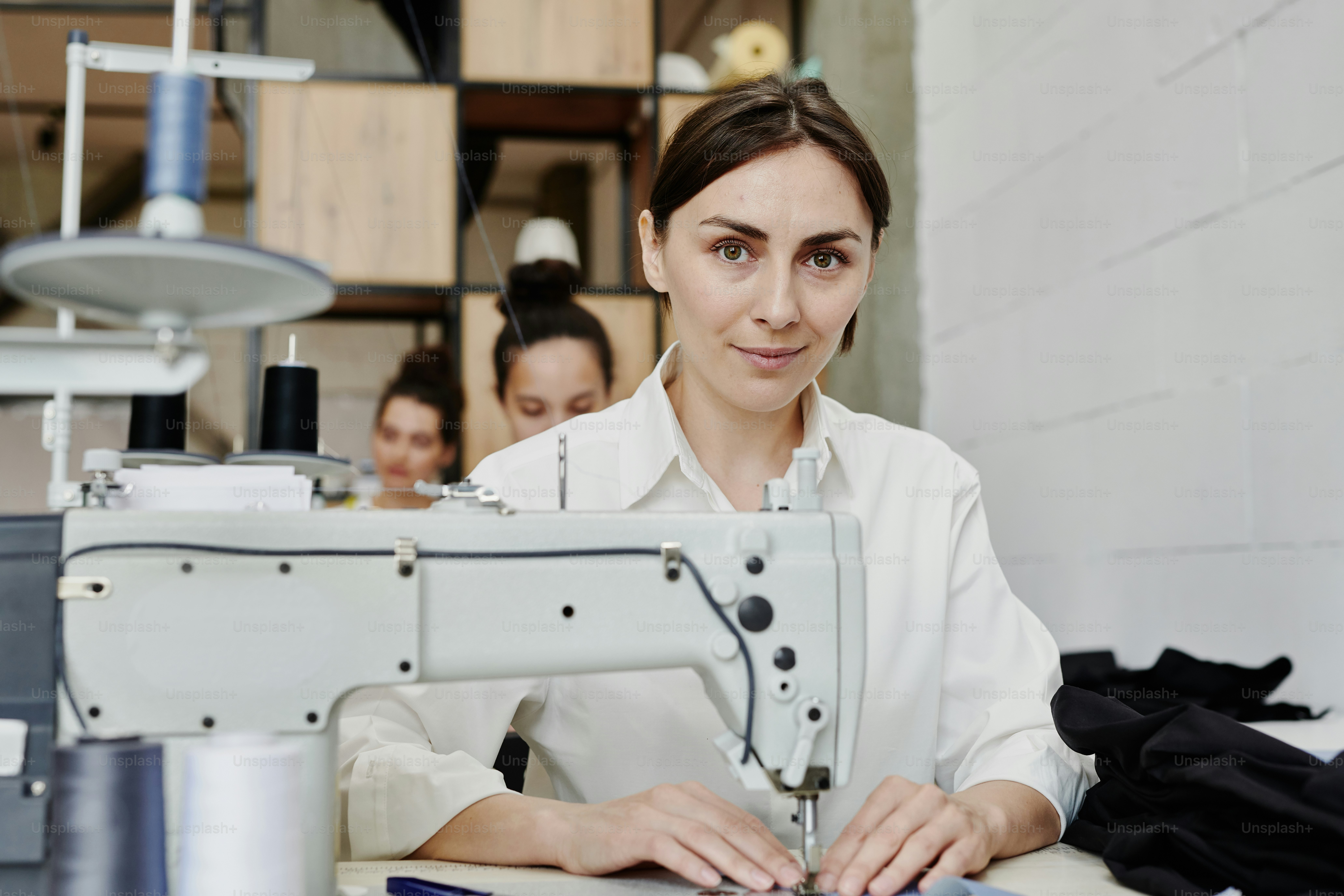 Giovane sarta contemporanea che ti guarda mentre sei seduta sul posto di lavoro con la macchina da cucire elettrica e fa o ripara vestiti
