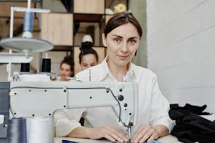 Junge zeitgenössische Näherin, die Sie ansieht, während Sie mit einer elektrischen Nähmaschine am Arbeitsplatz sitzen und Kleidung herstellen oder reparieren