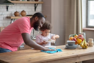 Dream team. Tall dark-skinned man in a pink tshirt wiping the table and smiling, his daughter wiping the plate