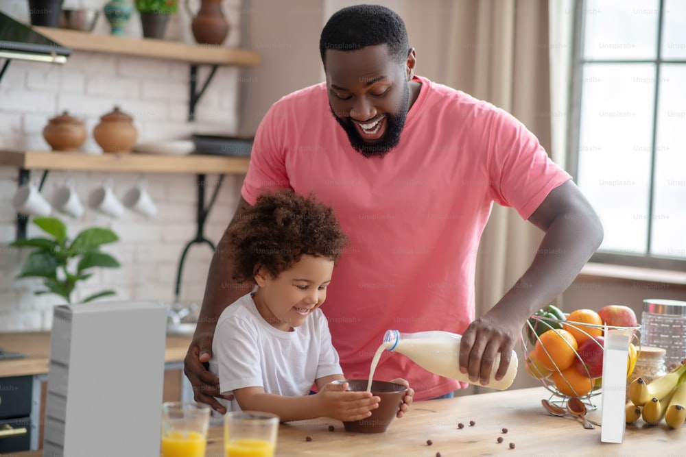 Getting breakfast ready, Black bearded man standing and pouring milk in his daughters plate