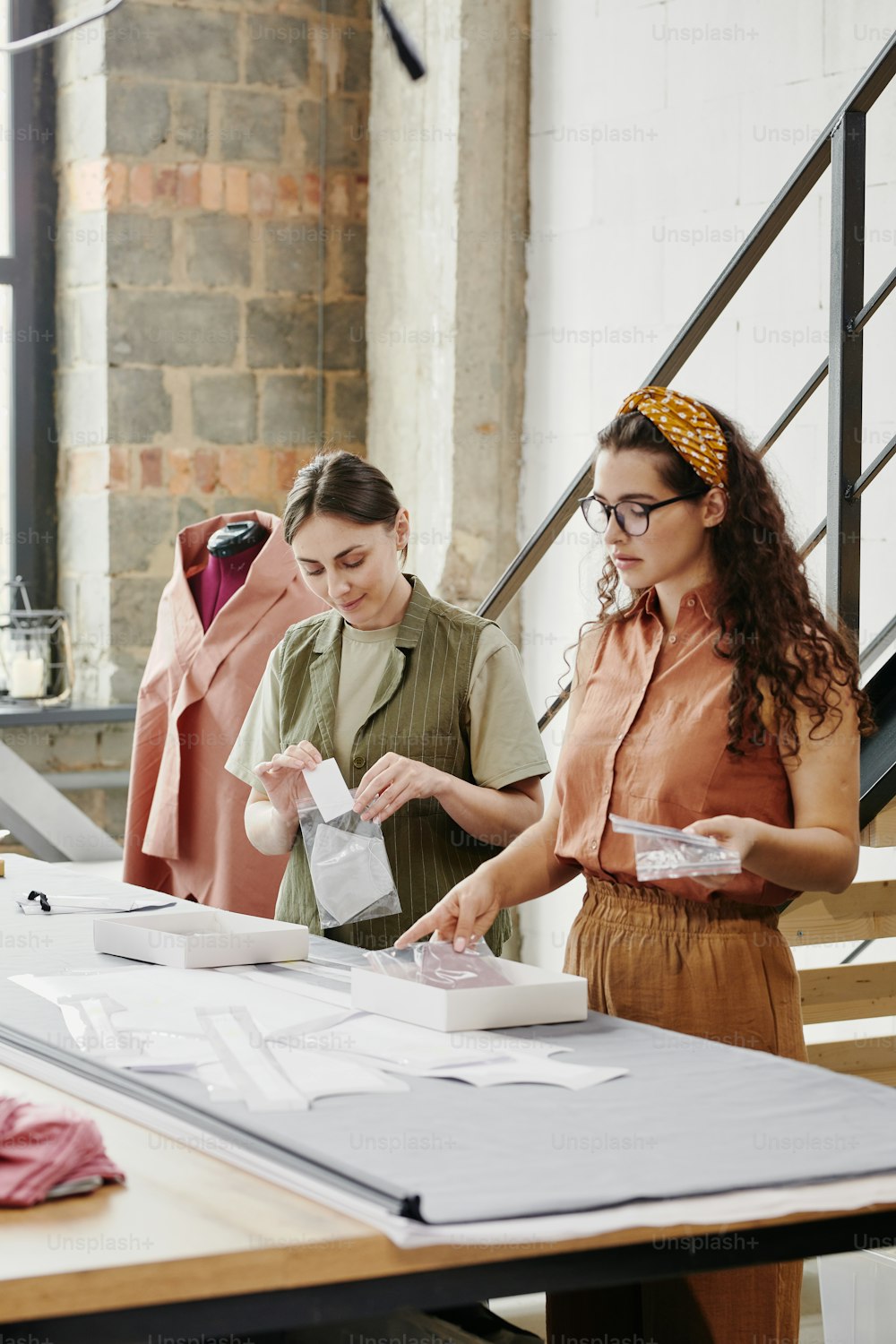 Two young female tailors in casualwear packing new shoulder pads for coats, jackets and dresses while standing by table in studio