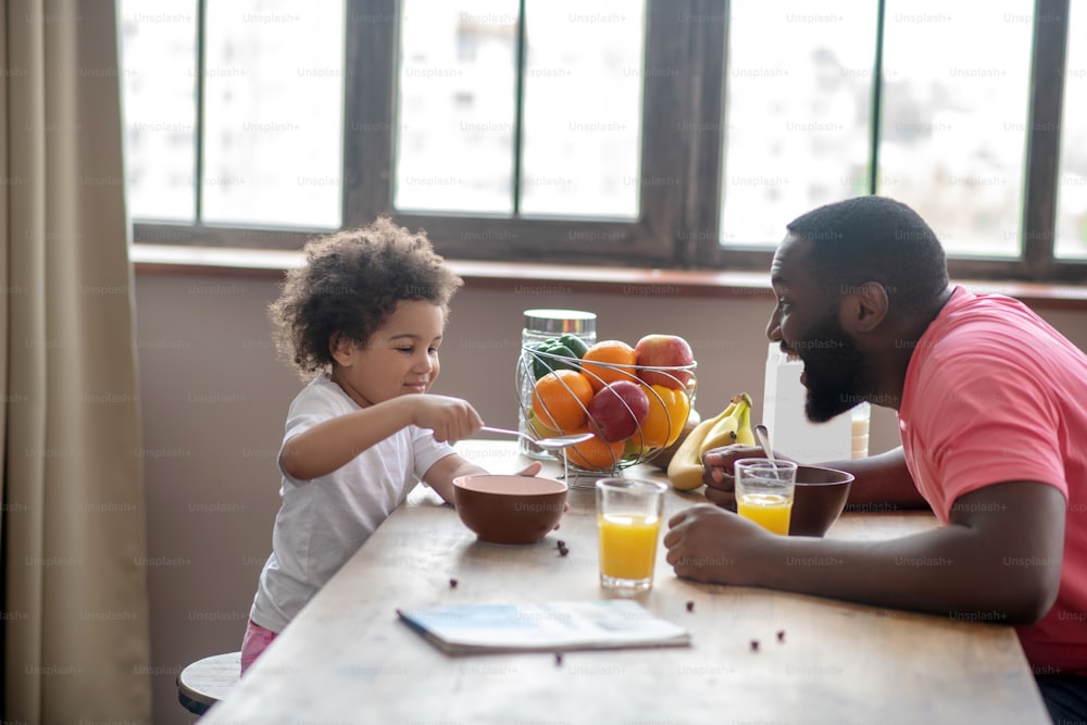 Funny breakfast.African american man in a pink tshirt entertaining his daughter while she eating