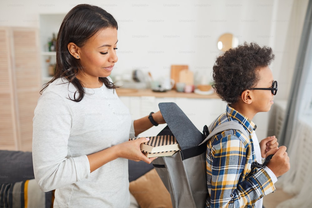 Side view portrait of smiling African-American woman putting books into backpack while sending son to school