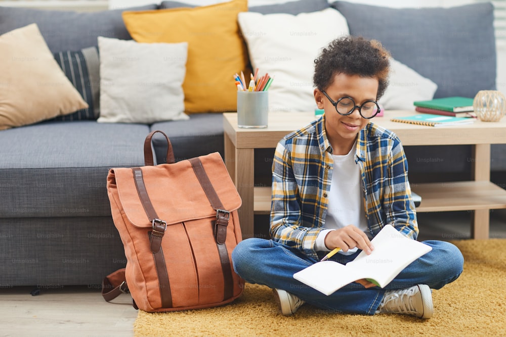 Full length portrait of cute African-American boy writing in notebook while sitting on floor at home, copy space