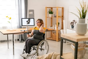 Cheerful disabled young female blogger in glasses sitting in wheelchair and streaming video at home