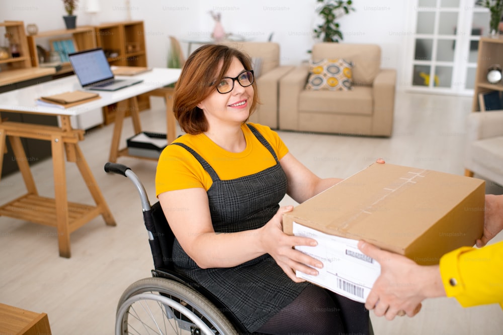 Entregador irreconhecível dando pacote para mulher com deficiência satisfeita em cadeira de rodas