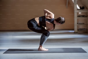 Beautiful brunette in active wear standing in Revolved Chair posture. Yoga studio interior.