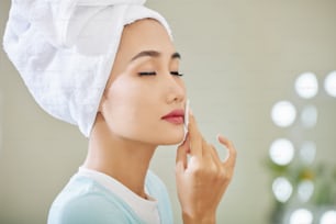 Hermosa joven asiática cerrando los ojos al limpiarse el lápiz labial con agua micelar en un disco de algodón