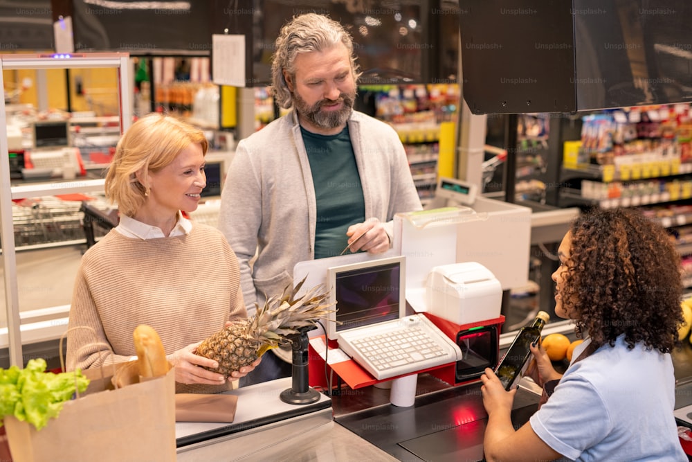 Mature couple standing by cashier counter in supermarket while young mixed-race woman scanning food products they bought
